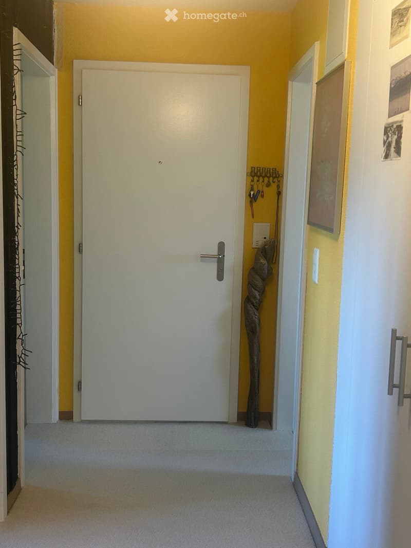 A narrow hallway with light yellow walls, carpeted floor, and white doors. A decorative wooden sculpture is placed near the wall on the right side.