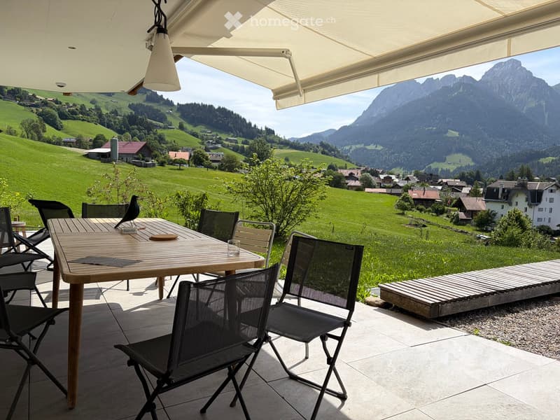 Balcony with a wooden table, chairs, and a bird figurine, overlooking a scenic mountain and valley landscape.