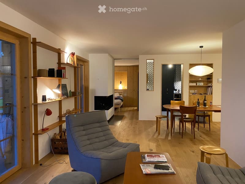 Modern, open-plan living and dining area with wooden flooring, a grey sofa, coffee table, dining table with chairs, shelves, and a small bedroom visible in the background.