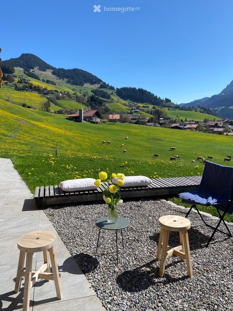 Outdoor seating area with wooden stools, a small table, a vase with yellow flowers, and a folding chair, overlooking a scenic valley with cows grazing and distant mountains.