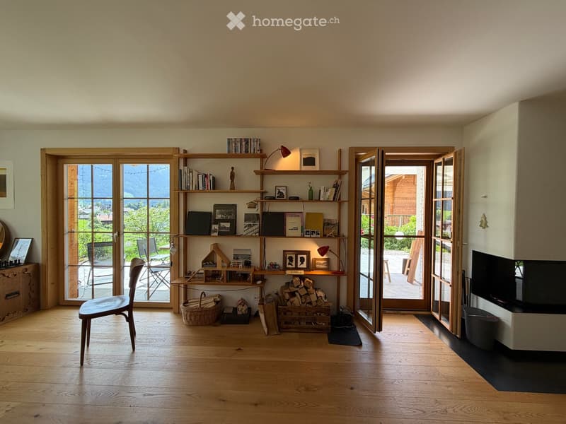 Bright living space with wooden flooring, large windows offering a scenic view, a wooden shelf filled with books and decorative items, a chair, a small table, and a television.