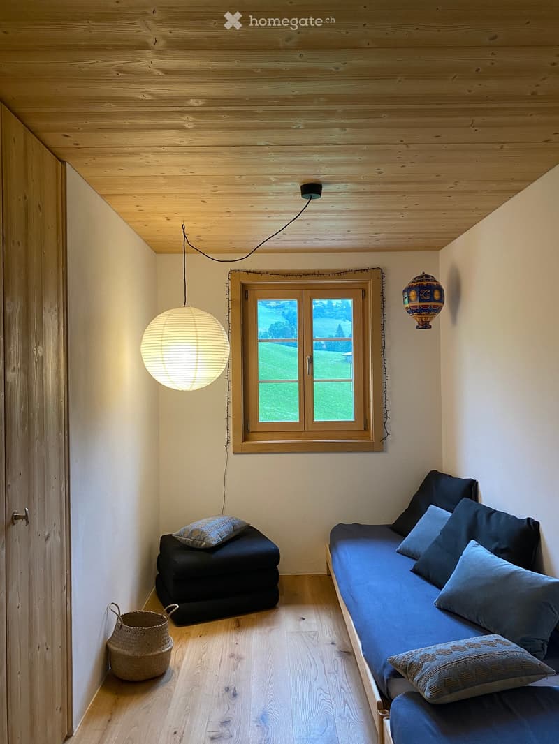 Cozy, compact room with wooden ceiling and floor, featuring a blue sofa, a black stool, and a wicker basket. A large window offers a view of a green landscape.
