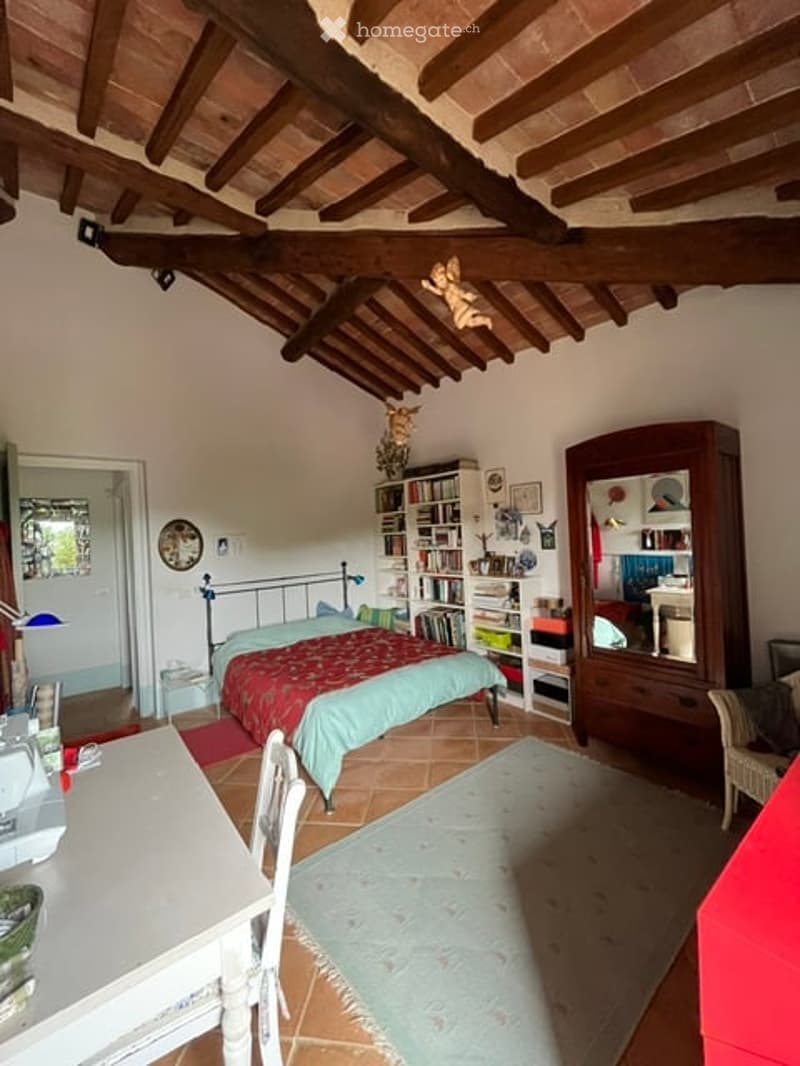 Cozy bedroom with exposed wooden beams, a bed with a red and green duvet, a bookshelf, a small table, and a mirror wardrobe.