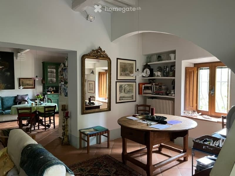 Cozy living and dining area with a mix of modern and classic decor. Features a large wooden dining table with chairs, a sofa, a mirror, a small bookshelf, and several picture frames on the walls.