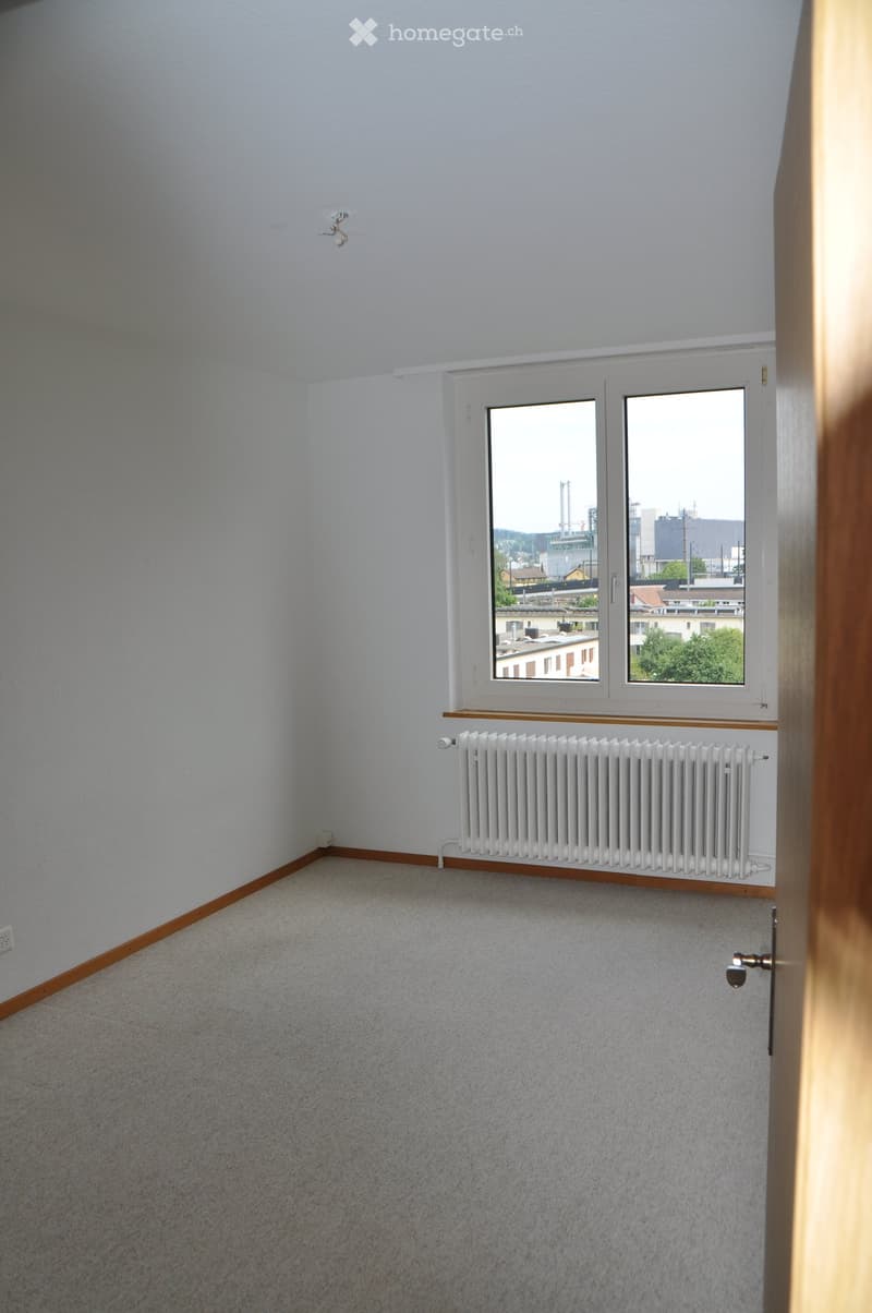 Empty room with light grey walls, carpeted floor, white radiator, and large window with a view of the city.