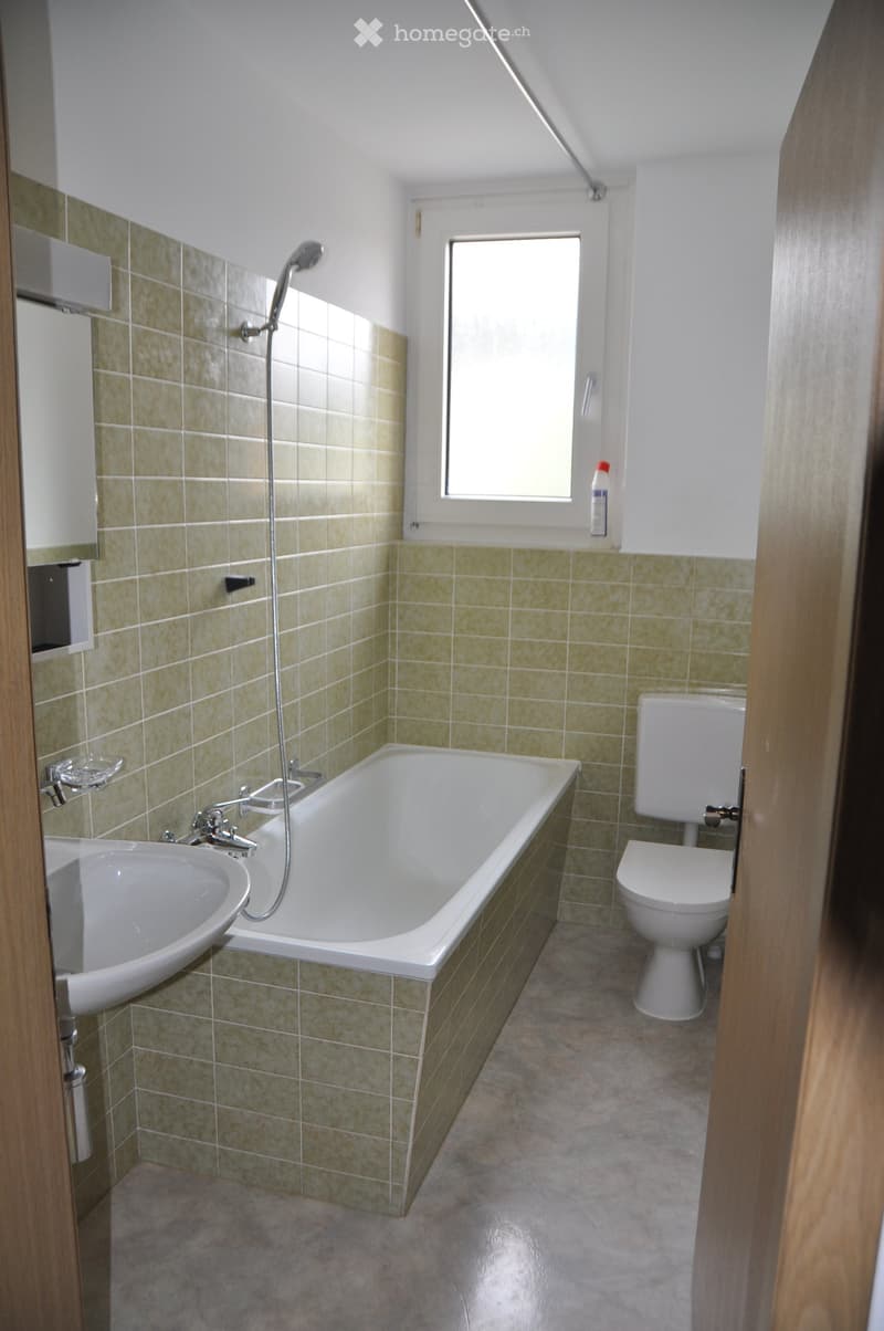 Bathroom with a bathtub, toilet, sink, and showerhead. Green tiled walls and a window providing natural light.