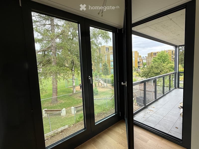 View from inside a room showing a modern balcony with large glass doors, wooden flooring, and a view of a green park area with trees and modern buildings.