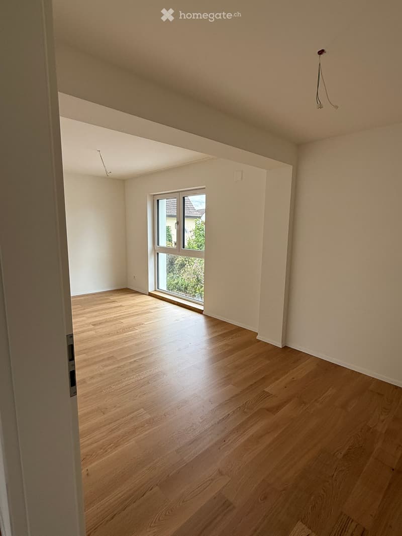 Empty room with light wooden flooring, white walls, and a large window with a view of greenery outside.