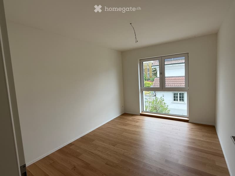 Empty room with wooden flooring and a large window showing a view of neighboring buildings and greenery.