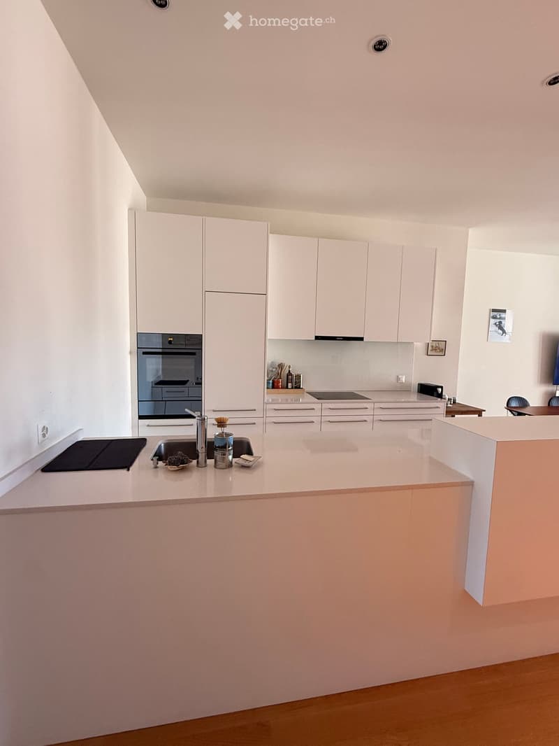 Modern kitchen with white cabinets, a central island, and stainless steel appliances including an oven and sink. The countertop features a few small items like a bowl and a bottle.