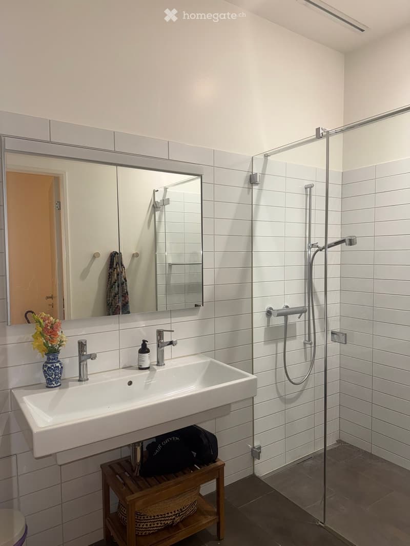Modern bathroom with white tiles, a large sink, a mirror, a shower, and a wooden shelf holding a basket and a hair dryer bag.