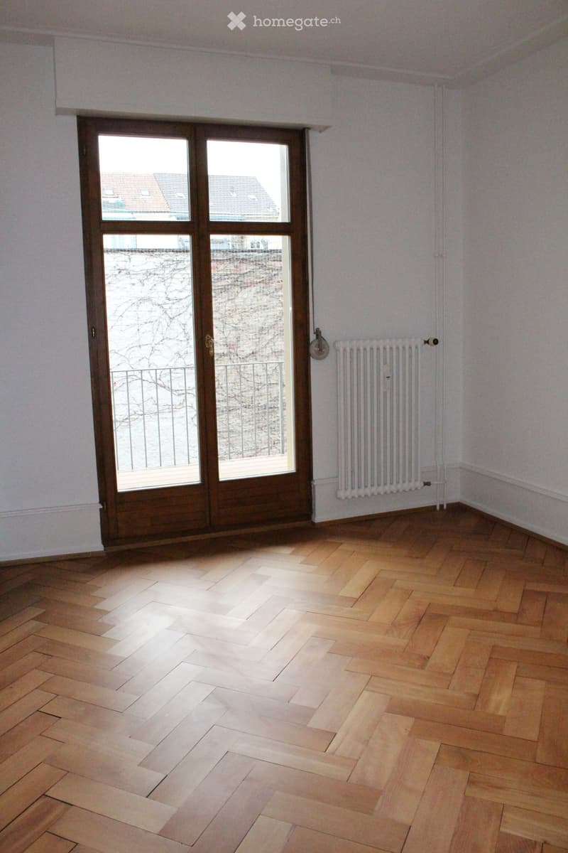 Empty room with wooden flooring, white walls, a radiator, and a large window with a view of a building and a balcony.
