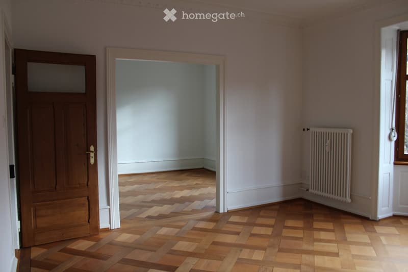 Empty room with wooden flooring, white walls, a radiator, and a doorway leading to another room.