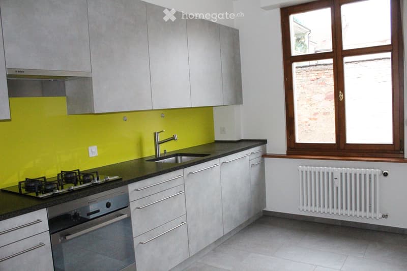 Modern kitchen with white cabinets, black countertops, a gas stove, an oven, a sink with a modern faucet, and a radiator under a large window.
