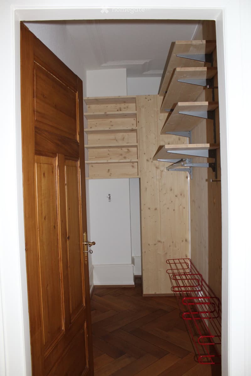 A narrow hallway with wooden flooring, white walls, and built-in wooden shelving units. A wooden door is open on the left, and a red wire basket is placed on the floor near the shelving units.