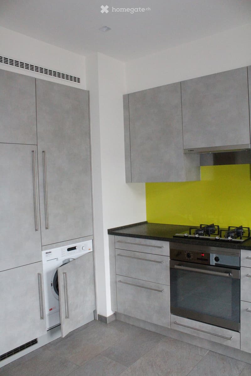 Modern kitchen with grey cabinets, a gas stove, an oven, and a private washing machine. The backsplash is a bright green color.