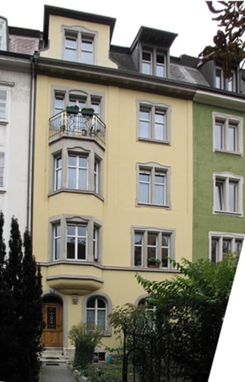 Multi-story residential building with yellow and green facade, multiple windows, and a small balcony on the second floor.