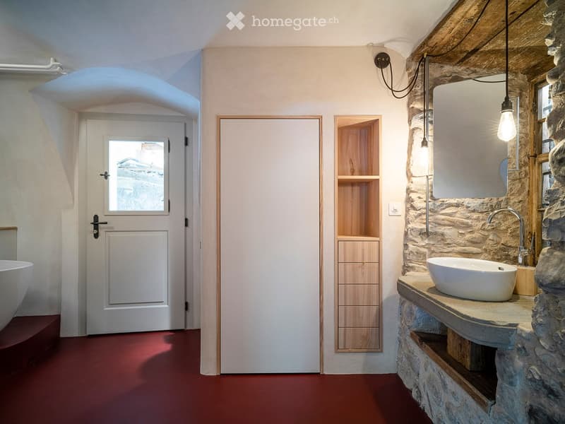 Modern bathroom with a stone sink, wooden cabinet, and a large mirror. The space features a red floor and a wooden door with a frosted glass panel.