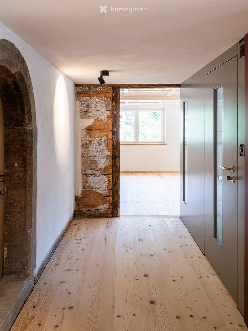 A narrow hallway with wooden flooring, white walls, and a partially exposed wooden beam. A door leads to another room with a window, and a modern light fixture is mounted on the ceiling.