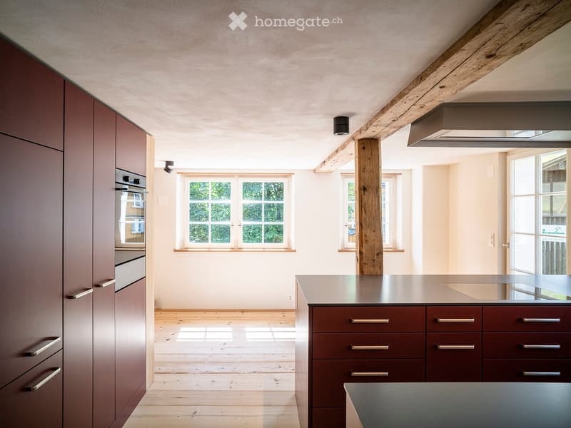 Modern kitchen with dark wooden cabinets, stainless steel countertop, and built-in oven. Natural light from large windows and exposed wooden beams.