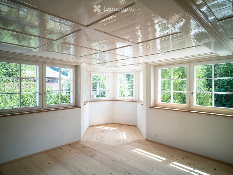Bright, empty room with wooden flooring, white walls, large windows, and a skylight ceiling allowing natural light to fill the space.