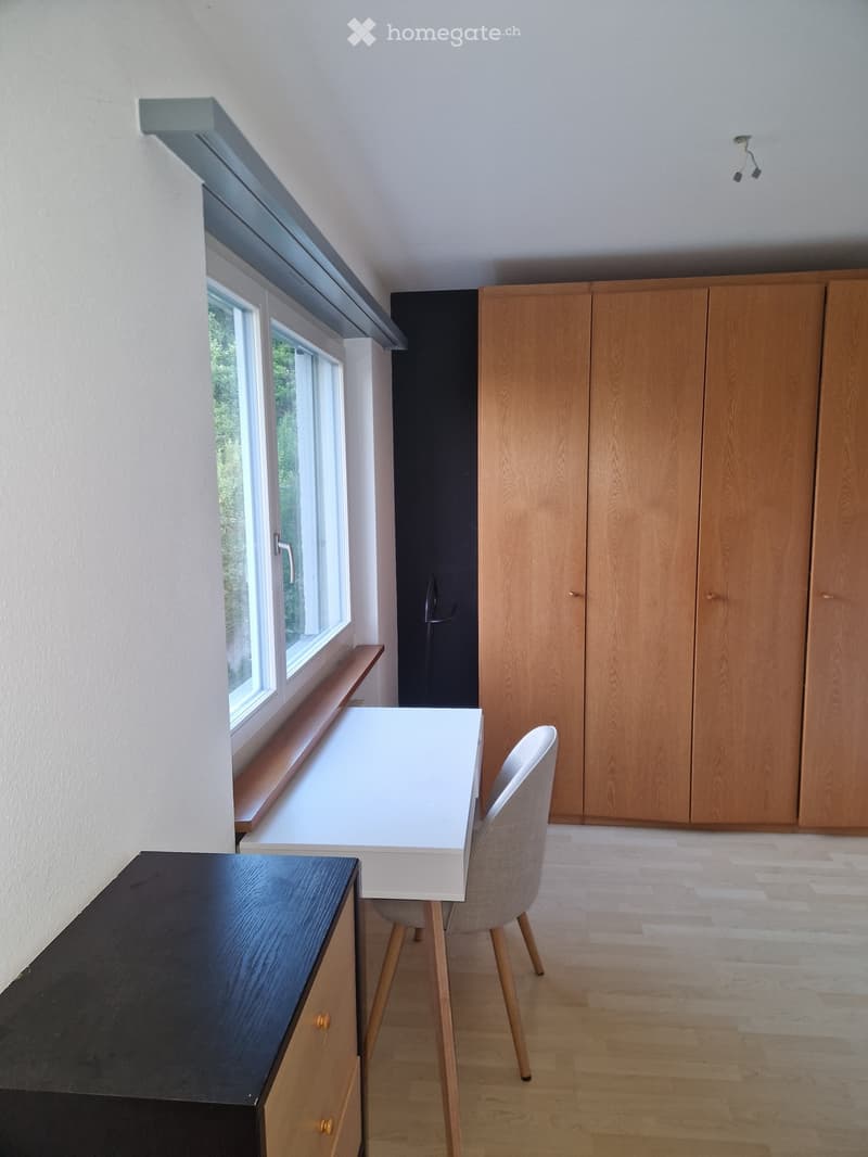 Bright room with wooden floor, white table, grey chair, and wooden cabinet. Large window with a view of greenery.