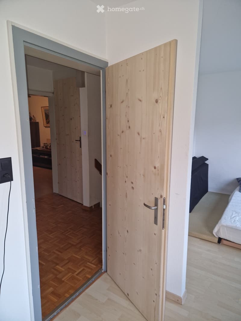 A narrow hallway with wooden flooring and white walls, featuring a closed wooden door and an open doorway leading to another room with visible furniture.