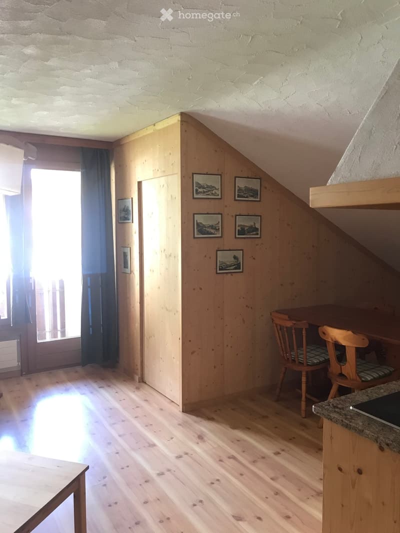 A small, cozy room with wooden floors, slanted wooden ceiling, and a window with dark curtains. There is a table with two chairs, a small counter with a granite top, and several framed pictures on the wall.