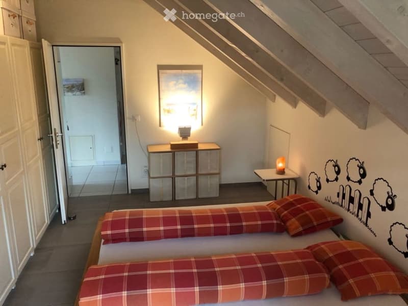 Cozy attic bedroom with slanted wooden ceiling, two single beds with red plaid bedding, nightstand with lamp, small table with chair, and wall-mounted picture frame.