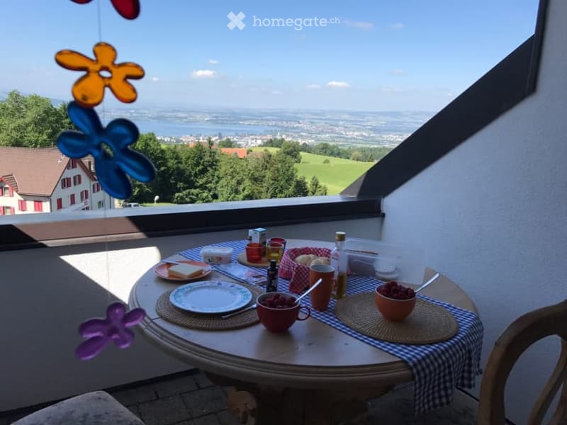 Balcony with a round table set for breakfast, overlooking a scenic landscape with a lake and distant town.
