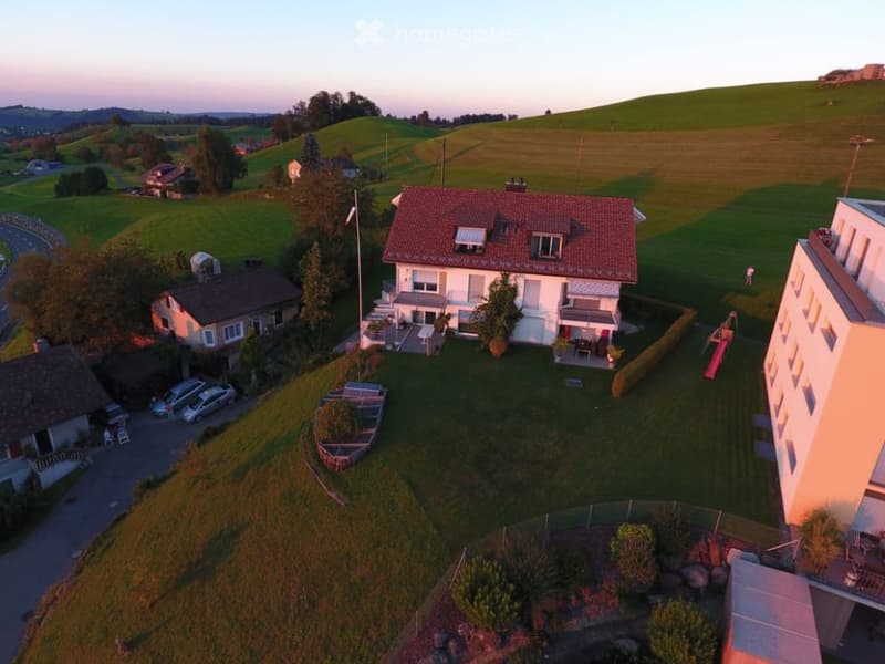 Aerial view of a two-story house with a red-tiled roof, surrounded by a well-maintained lawn, trees, and a small garden area. The house features multiple windows, a balcony, and a flagpole in front. Several parked cars are visible nearby, along with neigh