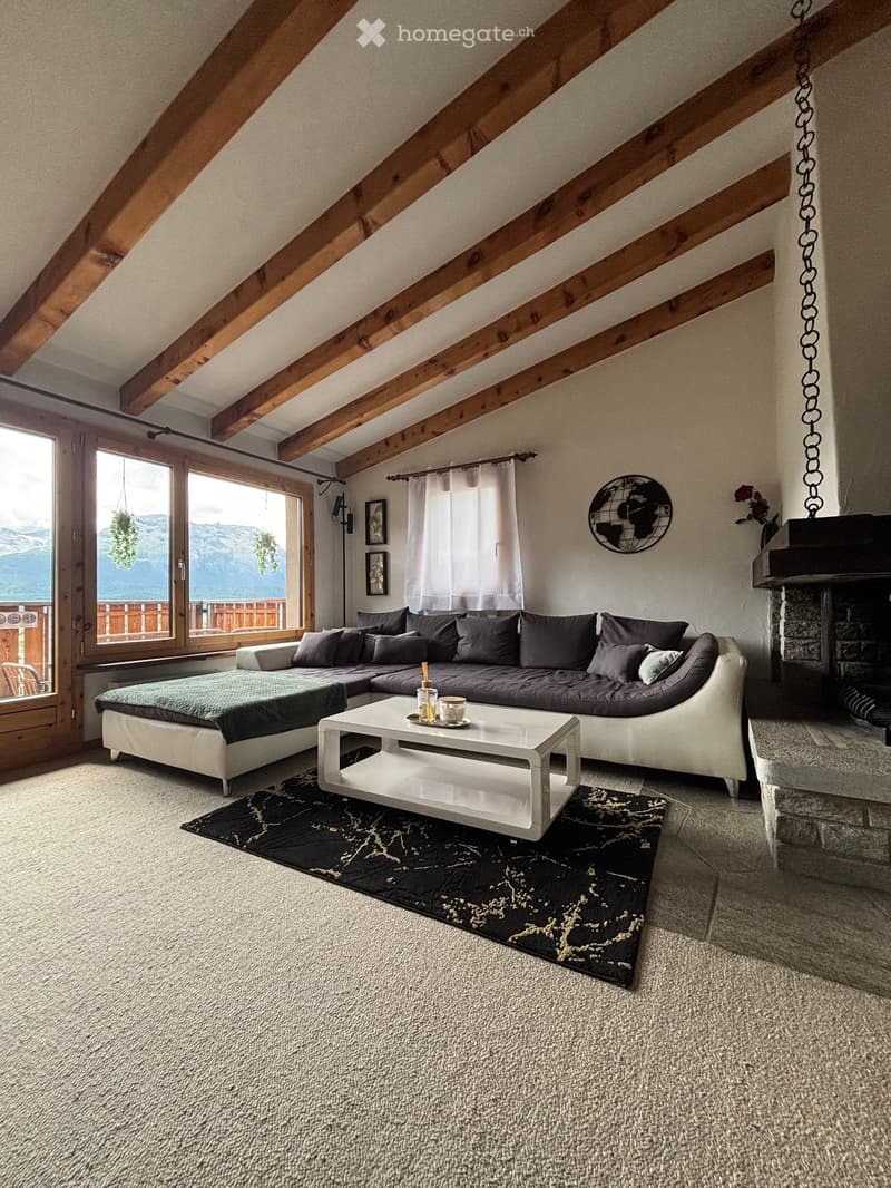 Modern living room with exposed wooden beams, large windows offering a mountain view, a sectional sofa, coffee table, fireplace, and decorative rug.