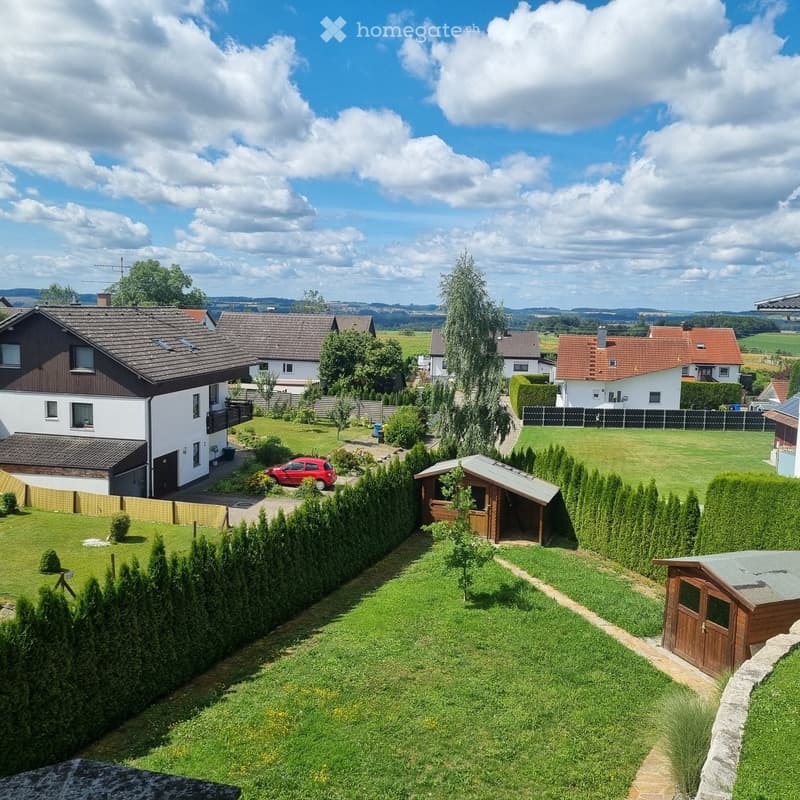A well-maintained garden with a lawn, trimmed hedges, a wooden shed, and a small wooden structure. A red car is parked near the house, and the surrounding area includes other houses and trees.