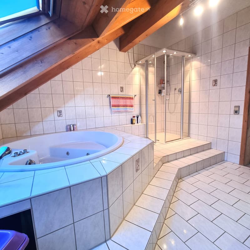 Modern bathroom with a large bathtub, glass-enclosed shower, tiled walls, and a skylight window. Features include a towel on a rack, a small shelf with toiletries, and a sleek faucet.