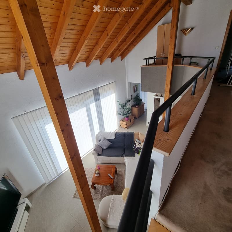 A modern living room with a grey sectional sofa, wooden coffee table, ottoman, and a small plant on a cabinet. The room features wooden beams on the ceiling and a staircase leading to an upper level.