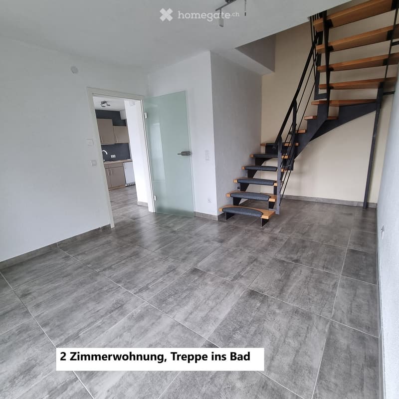 Empty room with grey tiled floor, staircase leading to another level, glass door to adjacent space, and visible kitchen area beyond.