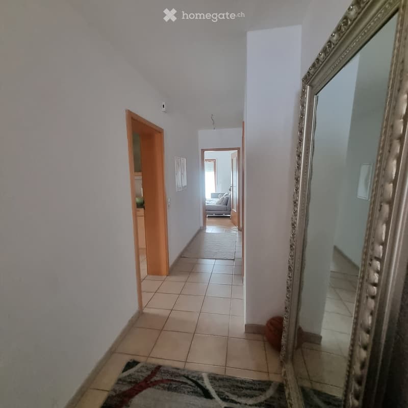 A clean, tiled hallway with light-colored walls, leading to an open door that reveals a living room area with a sofa and a rug.