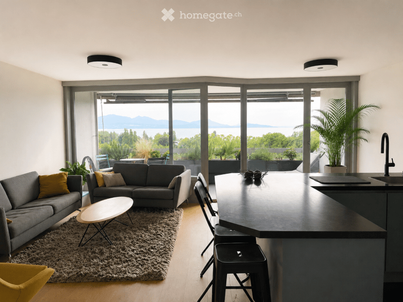 Modern open-plan living and dining area with large windows offering a scenic view, featuring a grey sectional sofa, coffee table, dining chairs, kitchen island, and plants.
