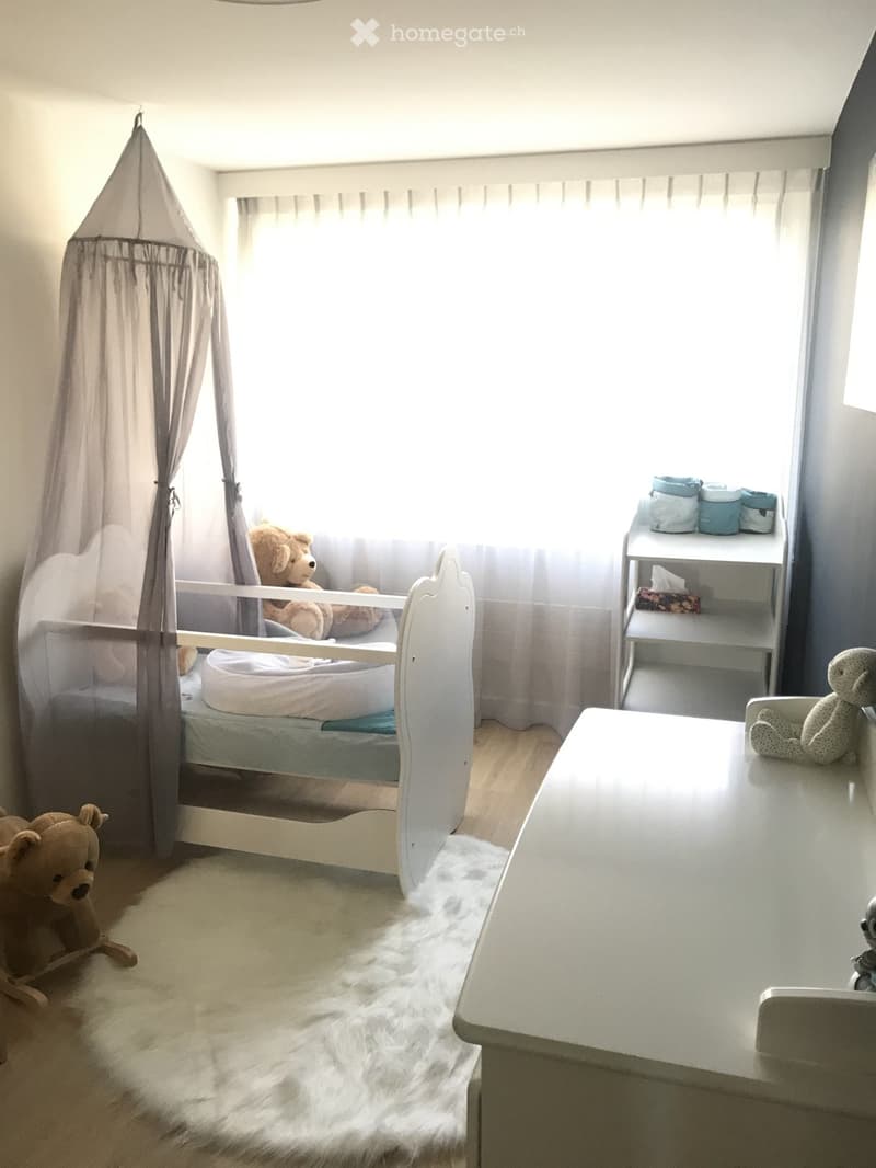 A well-lit children's bedroom with a white baby bed, a small table, a shelf with baby items, and several stuffed toys.