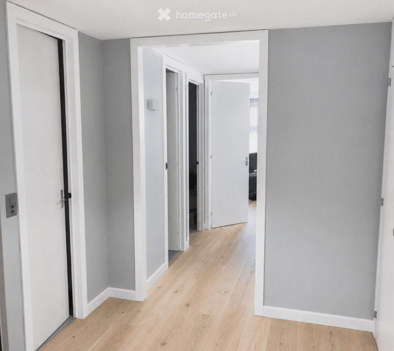 Empty hallway with light wooden flooring, grey walls, and white door frames. No furniture or equipment visible.