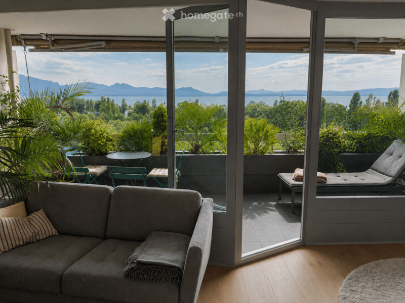 Living room with a grey sofa, wooden floor, large windows opening to a balcony with outdoor furniture and scenic mountain view.