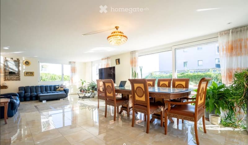 Bright living and dining area with large windows, a blue sofa, a wooden dining table with six chairs, a laptop, a television, and decorative items on the walls.