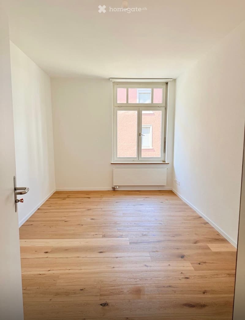 Empty room with light wooden flooring, white walls, and a single window with a radiator below it.