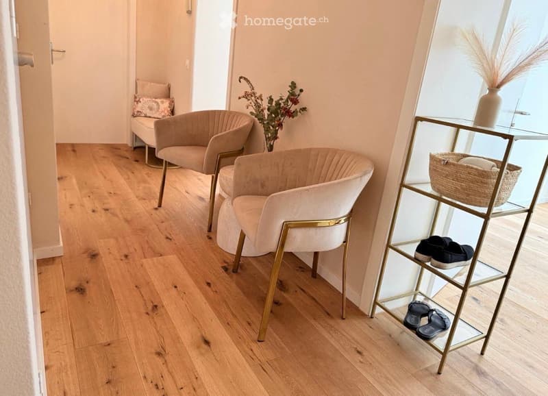 A small, well-lit hallway with light wooden flooring, two beige chairs, a glass shelf holding shoes and a basket, and a vase with dried plants.