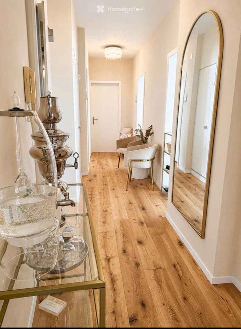 Elegant hallway with light wooden flooring, white walls, and a large round mirror on the right. A glass table with a coffee machine and glassware is visible on the left, and a small seating area with white chairs is seen in the background.