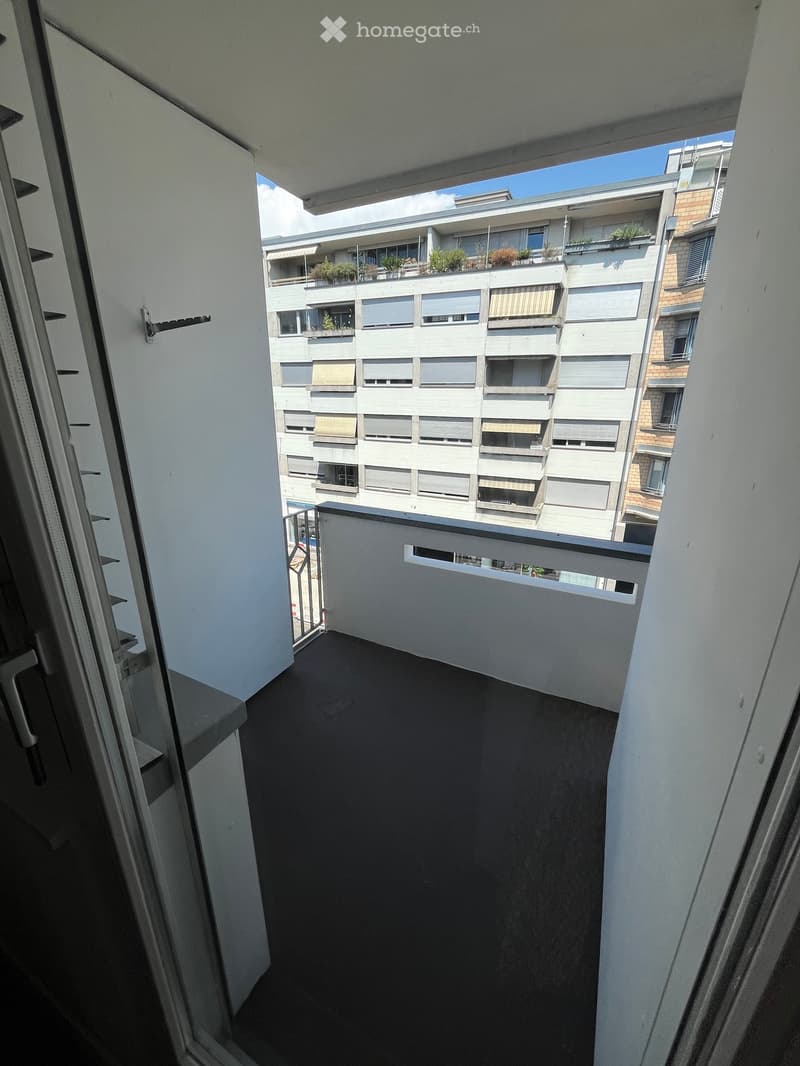 Balcony with dark flooring, white walls, and a view of a multi-story building with balconies and greenery on the rooftop.