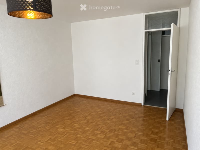Empty room with wooden herringbone flooring, white walls, and a hanging lamp. An open door leads to another room.