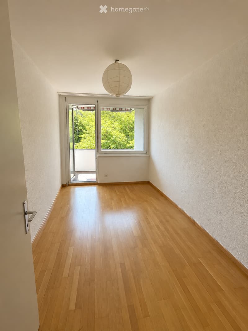 Empty room with wooden flooring, white walls, and a large window with a sliding door leading to a balcony. A single hanging light fixture is visible on the ceiling.