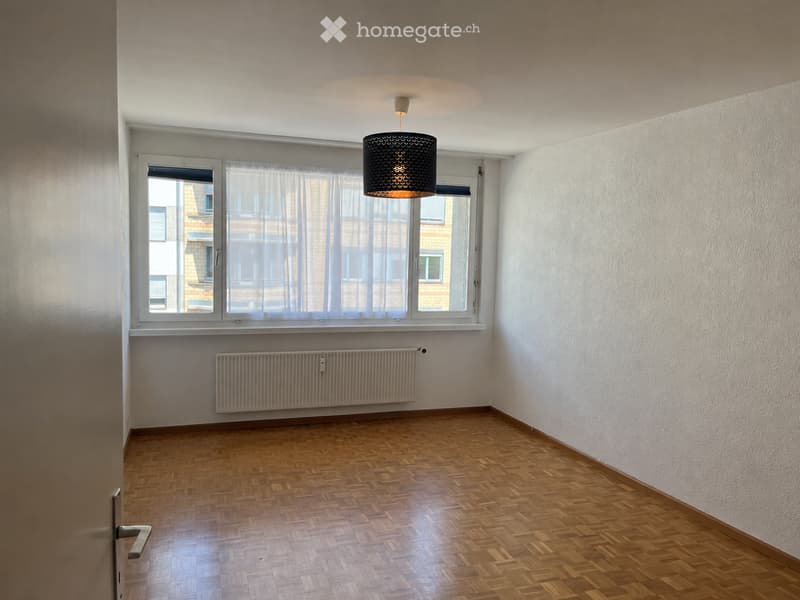 Empty room with wooden parquet flooring, white walls, large window with sheer curtains, radiator, and a hanging light fixture.