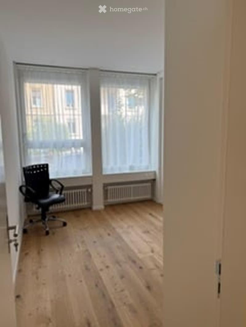 Minimalist room with light wooden flooring, large windows with white blinds, and a single black office chair.